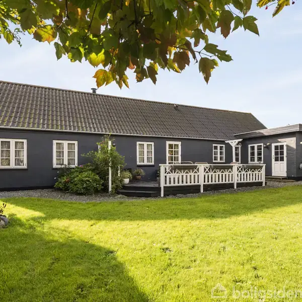 En sortmalet villa med hvide vinduer står i en solrig have med en grøn græsplæne. En træterrasse med rækværk ses foran huset.