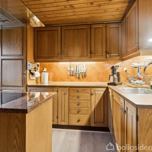 Køkken med træskabe og -loft, hvor knive hænger på væggen over en bordplade. En kaffemaskine og vask i nærheden. Hyggelig belysning skaber en varm atmosfære.