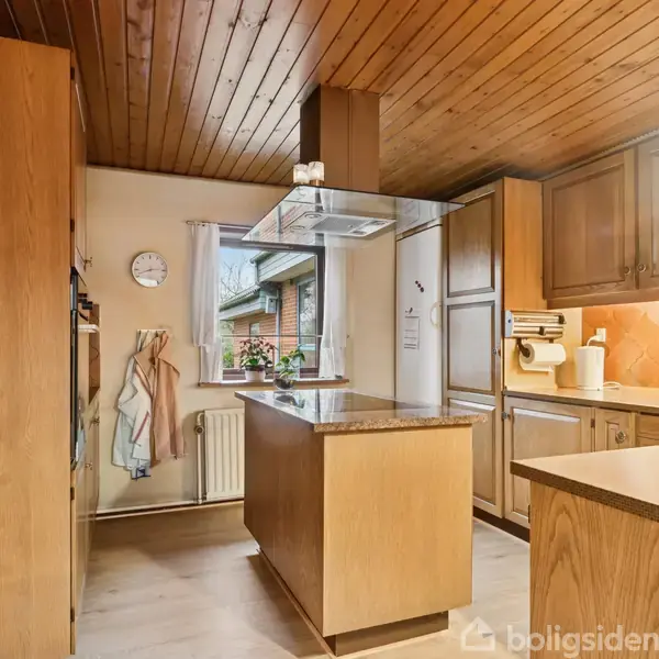 Et køkken med trækabinetter har en kogeø med emhætte. Vinreol er fyldt, og knive hænger ved væggen. Der er også en vask og køkkenudstyr på bordpladen.