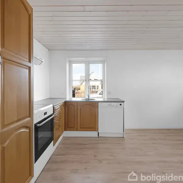 Køkken med trækøkkenskabe og moderne hvidevarer. Køleskab, ovn og opvaskemaskine indbygget. Store vinduer slipper lys ind fra udendørs område. Lyse trægulve og hvidt loft skaber en lys atmosfære.