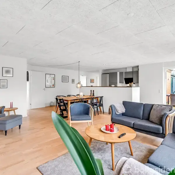 En lys stue indeholder en sofagruppe med sofabord og lamper. I baggrunden er der et spisebord, og et åbent køkken er synligt. Indretning med planter og billeder på væggen.