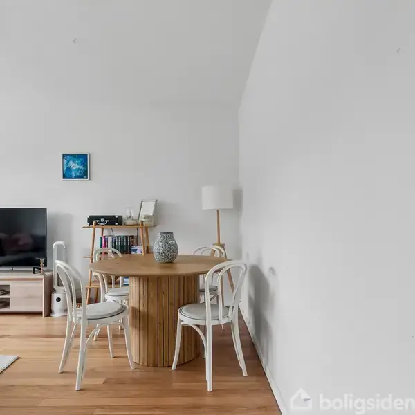Et rundt træbord med fire hvide stole står i et lyst rum med parketgulv. I baggrunden ses et TV på et lavt bord samt en bogreol med lamper og dekorationer.