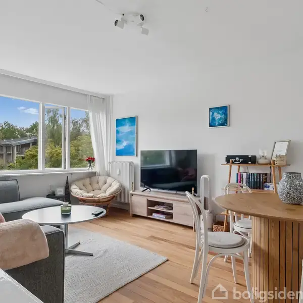 En moderne stue med en stor sofa og rundt sofabord, en fladskærms-TV på et skab, træspisebord med hvide stole, en lænestol, lyse vægge, og stort vindue med udsigt til træer.