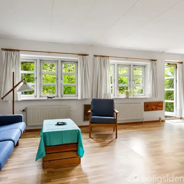 En lys stue med trægulv og hvide vægge har en blå sofa og lænestol omkring et bord med en turkis dug. Store vinduer med hvide gardiner udsigt til grønne omgivelser.