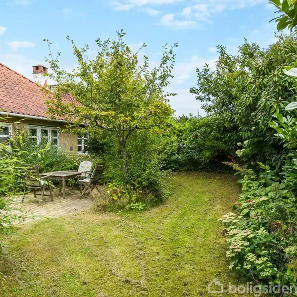Hus med rødt tegltag og flere vinduer, omgivet af tæt vegetation. I forgrunden et havebord og stole på en terrasse med græsplæne og buske omkring.
