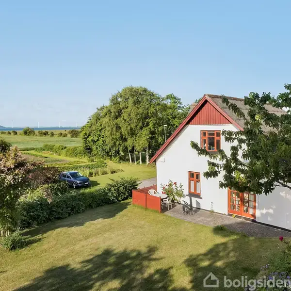 En hvid, to-etagers villa står stille i en velplejet have med græsplæne og træer. I baggrunden ses åbne marker og en blå himmel. En parkeret bil er synlig.