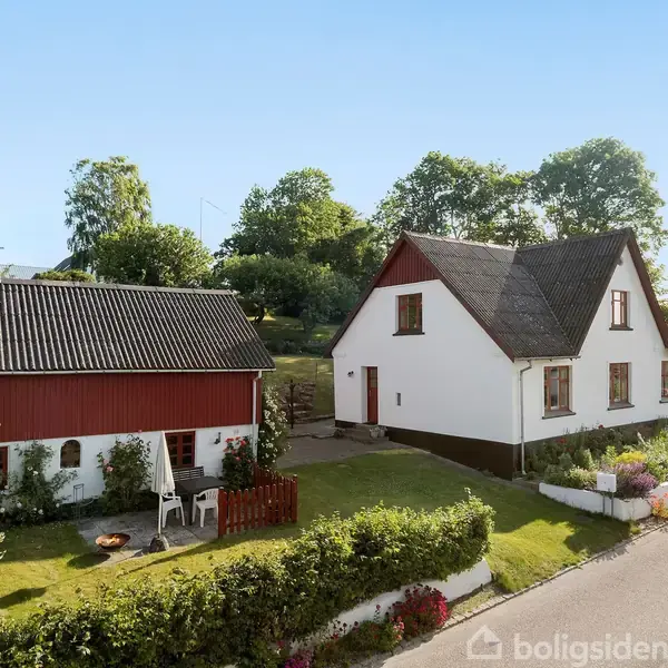 To huse står side om side; det ene er hvidt med mørkt tag, det andet rødt med sort tag. Foran ligger en have med blomster og en havegang.