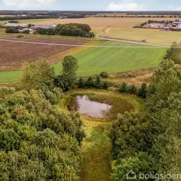 Et stort hus omgivet af træer og marker set fra luften; en gårdsplads og flere udhuse er synlige. Landskabet er præget af åbne marker i baggrunden.