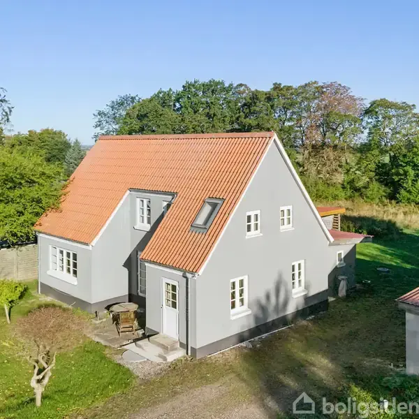 Et gråt hus med en rød tagstenstag står i en have med græsplæne og træer. Huset har flere vinduer, omgivet af grøn vegetation og blå himmel.
