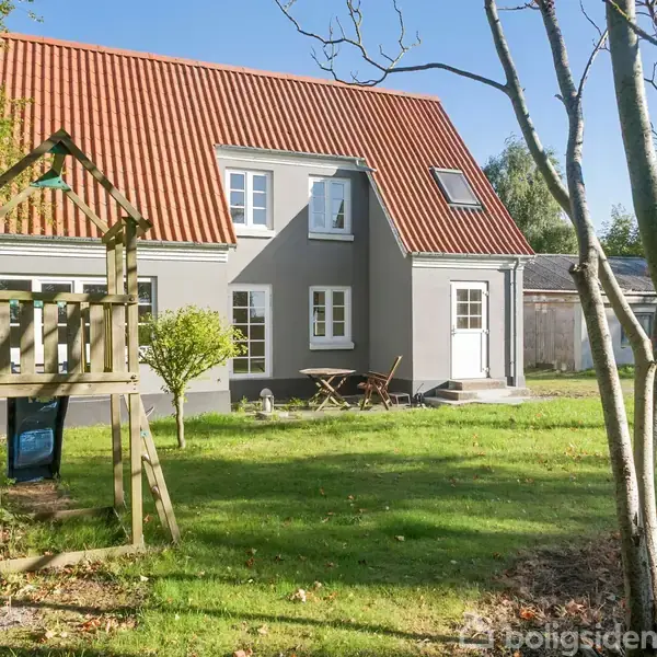 Grå hus med rødt tag står i en solrig have. En trælegeplads med skydestige og hængebro er placeret i forgrunden. Træer omgiver området.