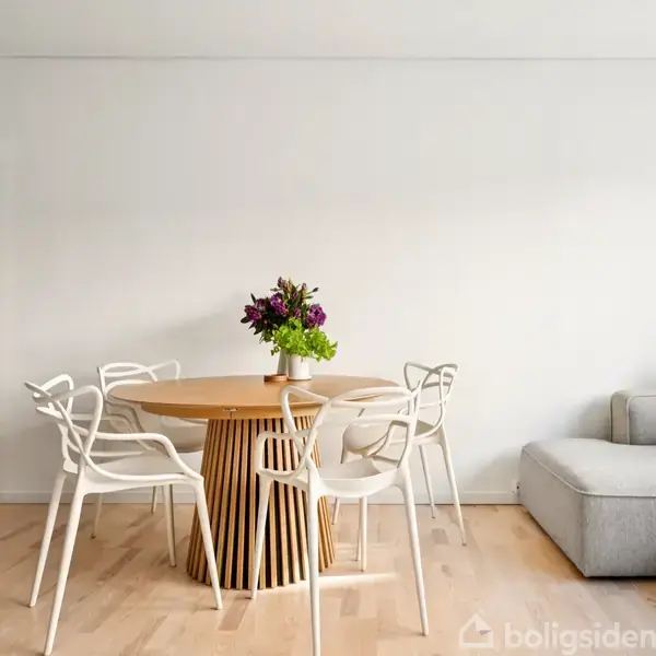 Et rundt træbord med en buket blomster står i en lys stue omgivet af moderne, hvide stole og en grå sofa. En åben dør ses i baggrunden.