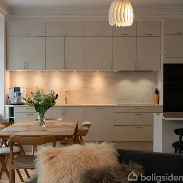 En moderne køkken-spisestue med hvidt køkkenskab, spisebord med blomster i vase og lamper hængende fra loftet. Store vinduer giver naturligt lys. Ingen tekst ses i billedet.