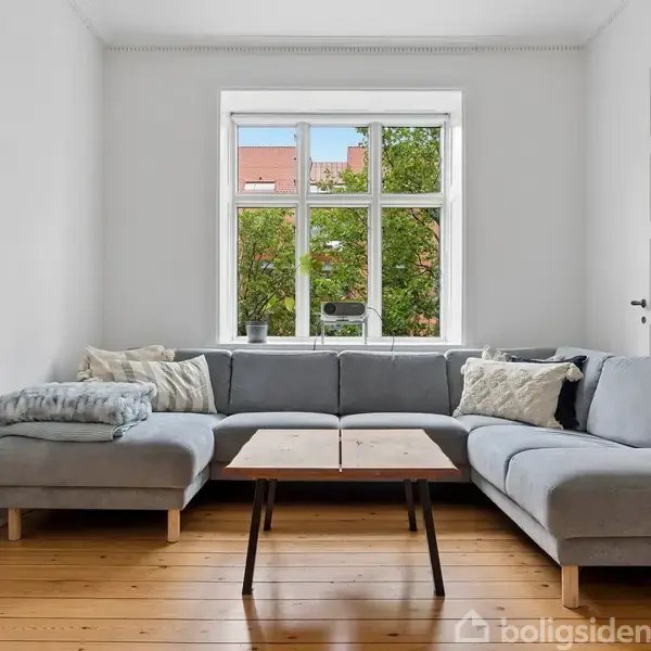 Grå sofa står ved et lyst vindue med trærammer, omgivet af hvide vægge og trægulv. Sofabordet foran sofaen er enkelt. Udsigt til grønne træer gennem vinduet.