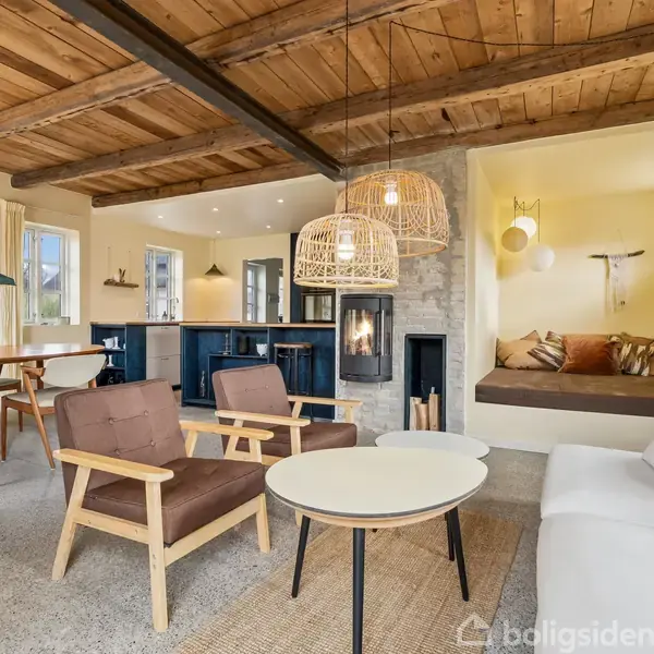 Stue med træmøbler; sofa og lænestol omkring runde borde. Pejs ved stenmur. Åben køkken med spisebord. Træloft og moderne lamper skaber hyggelig atmosfære.