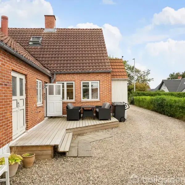 Rødstenshus med terrasse, hvor havemøbler står opstillet. Grusvej løber langs huset, omkranset af hæk, mens en bænk og plantekrukker dekorerer området. Himmel oplyst og let skyet.