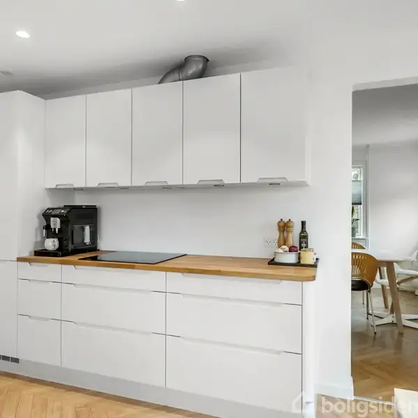 Et moderne køkken med hvide skabe og træbordplade. En kaffemaskine og køkkenredskaber står på bordet. I baggrunden ses et spiseområde med naturligt lys.