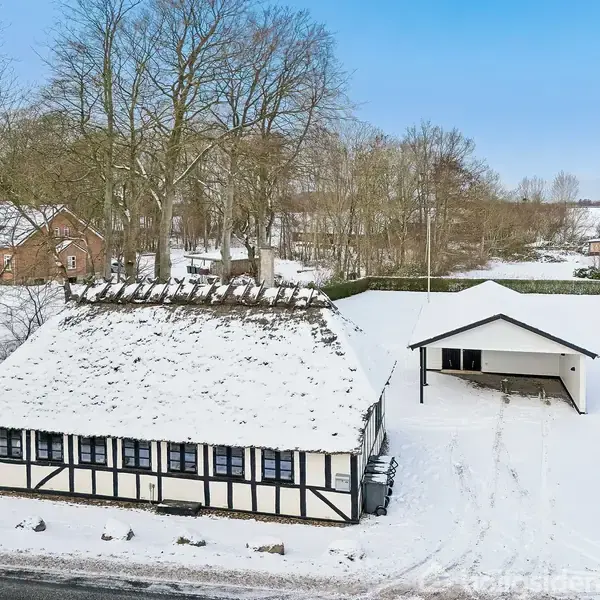 Et bindingsværkshus dækket af sne står ved siden af en lille garage. Omgivet af snedækkede marker og træer, skaber det en idyllisk vinterlandskab.