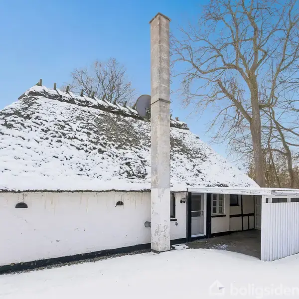 Et bindingsværkshus med stråtag er dækket af sne og står roligt i et skovområde om vinteren. En skorsten rager op, og et lille skur er ved siden af huset.