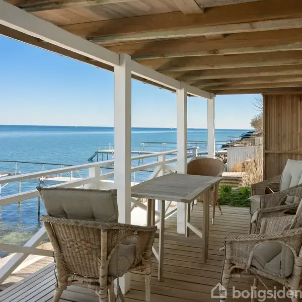 En overdækket terrasse med seks flettede stole og et bord, der vender ud mod havet. Solrigt vejr og en rolig kystlinje skaber en afslappende atmosfære.