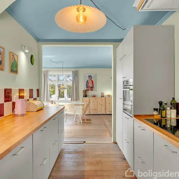 Køkken med lyse træbordplader, hvide skabe, og rød-hvide fliser. Rummet fører ind til en hyggelig spisestue med lyseblåt loft, hvidt spisebord, og stort vindue.