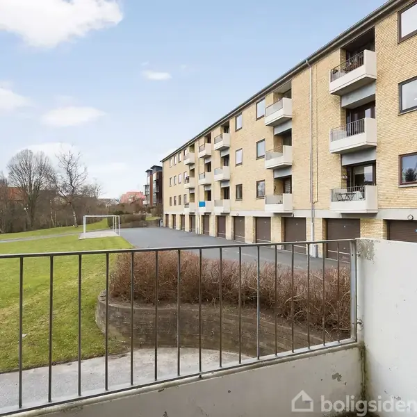 En altan med metalrækværk hænger ud fra en bygning, der er lavet af mursten med flere vinduer. Handlingen viser udsigt til en grøn græsplæne med en fodboldmålramme.