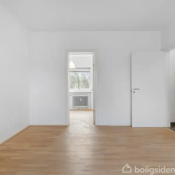 Et tomt rum med trægulv, en åben dør i midten fører til et andet værelse, og en lukket dør til højre. Vinduet i baggrunden viser natur udenfor.