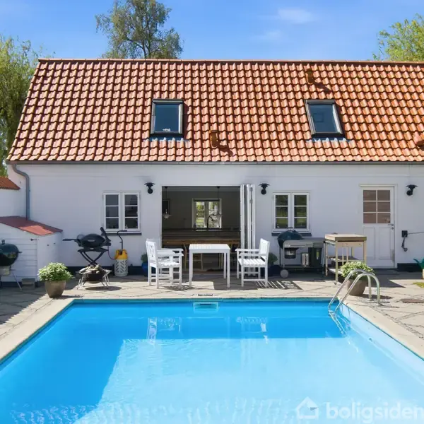 Hvidt hus med rødt tegltag, har en udendørs swimmingpool og terrasse. Solrige omgivelser med havemøbler og grill ved poolkanten. Grønne træer i baggrund.