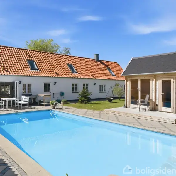 En udendørs swimmingpool med klart vand ligger foran et hvidt hus med rødt tegltag. Til højre står en lille pavillon ved poolkanten, omgivet af grønne planter og græsplæner.