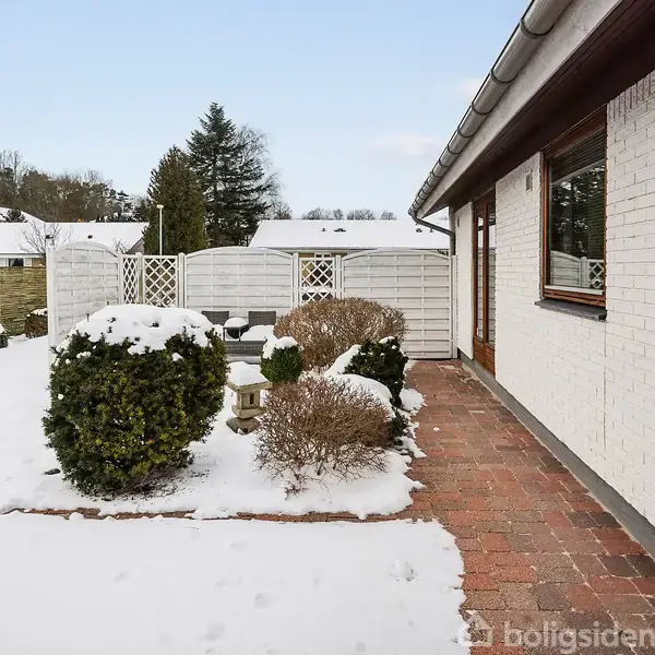 Hus med hvid murstensfacade, beliggende ved en snedækket have med buske og flisesti. I baggrunden ses et hegn og flere træer, hvilket skaber en rolig omgivelse.