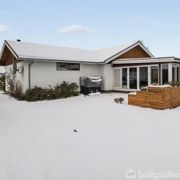 Et hvidmalet hus med en stor glasveranda står i en snedækket have. Der er hæk på venstre side og en opbygget plantekasse foran verandaen.
