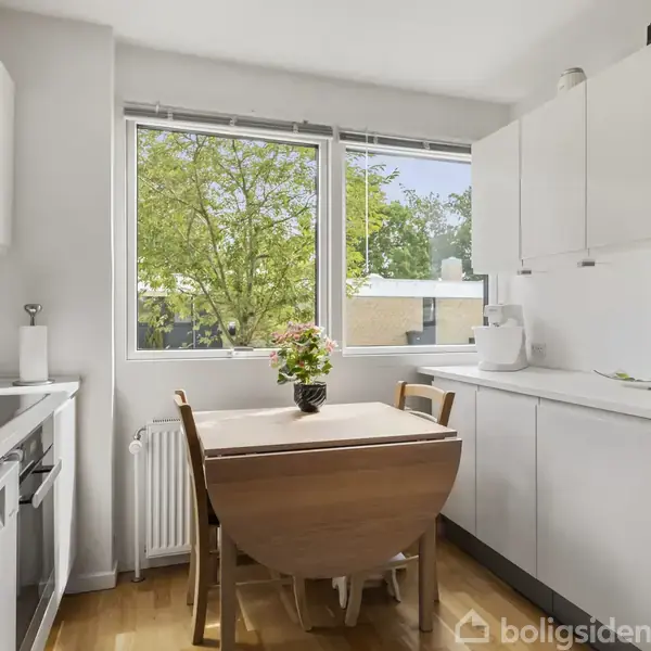 Et lyst køkken med en hvid bordplade og skabe. Et lille træbord med blomster står i midten. Et vindue viser grønne træer udenfor.