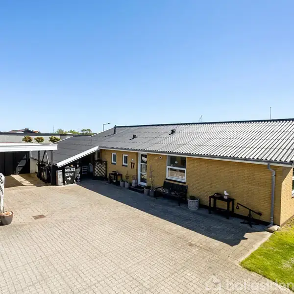 Et gulstenshus med skråt tag står stille på en flisebelagt gårdsplads. I baggrunden ses en carport og en gade med lygtepæle, omgivet af grønne områder under en klar blå himmel.
