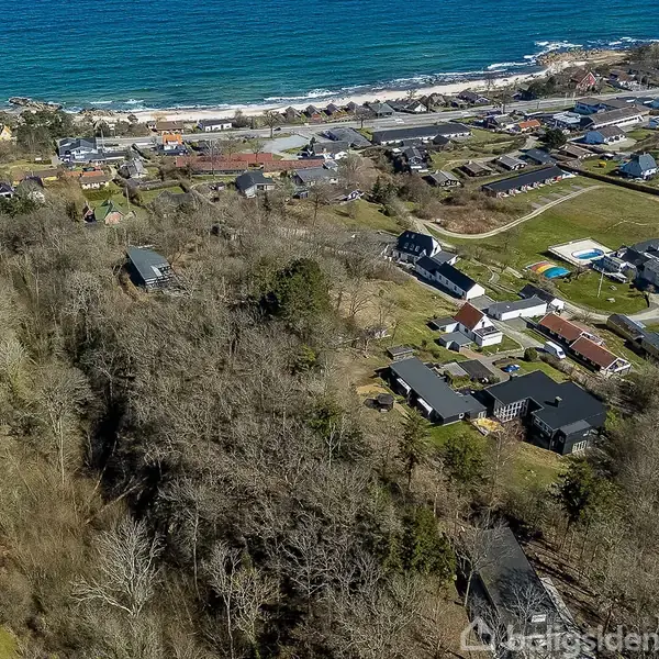 Huse ligger spredt i et skovområde tæt ved kysten. Havet strækker sig i baggrunden, omgivet af en række boliger og græsklædte områder.