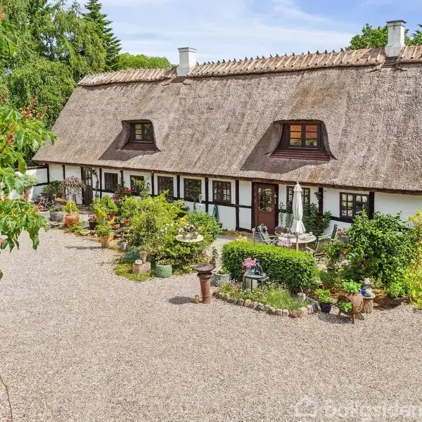 Et gammeldags bindingsværkshus med stråtag står i en grusbelagt gårdsplads. Der er frodige planter og blomsterkrukker langs facaden, omgivet af grønt løv og træer i baggrunden.
