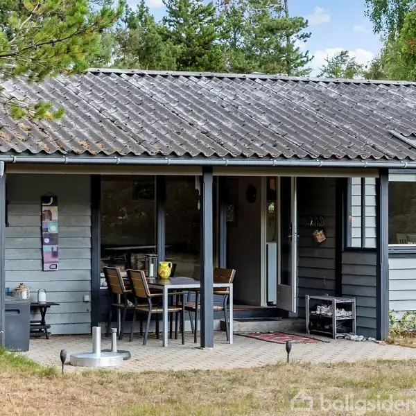 Et gråt træhus står i en grøn have. Bord og stole er placeret foran huset. Skoven danner baggrund med blå himmel og få skyer.