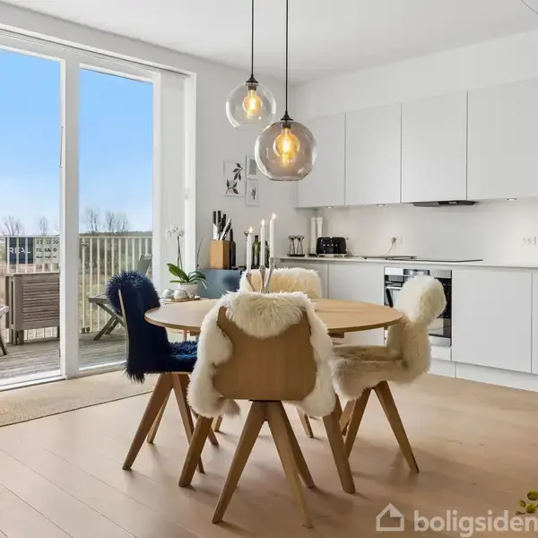 Spisebord med fire stole dækket af pels, står i et køkken-alrum med hvide skabe. Store vinduer og åben altandør giver udsigt til en balkon. Dekorative lamper hænger over bordet.