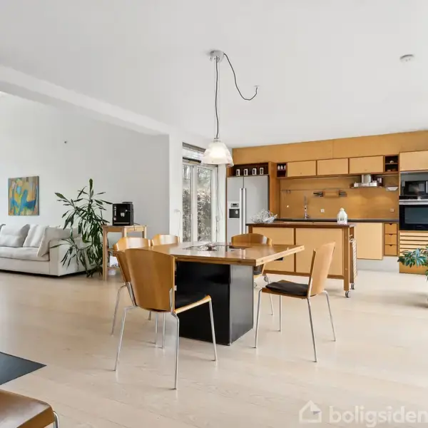 En moderne stue med træmøbler og et spisebord i midten. En sofa står i hjørnet ved en plante, og der er et åbent køkken i baggrunden.