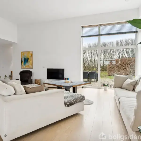 En lys stue med hvide sofaer står i en moderne indretning. Et stort vindue bagved giver udsigt til en have. Et fjernsyn er placeret på et lavt bord.