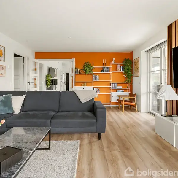Sofaen står i stuen med en mørkeblå farve. Den vender mod et stort TV på væggen. Bagved ses en orange bogreol fyldt med bøger og planter. Væggene har forskellige kunstbilleder. En glasdør leder ud til et lyst område. Trægulv dækker rummet, og et sofabord står foran sofaen med en vase med tulipaner.