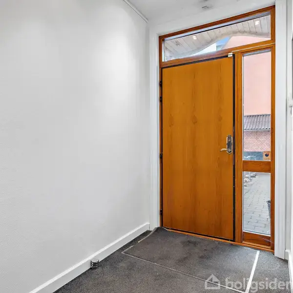 En trædør står åben i en lys, hvidmalet gang med et enkel gråt gulvtæppe. Udenfor ses en rød bygning og flisebelagt område.