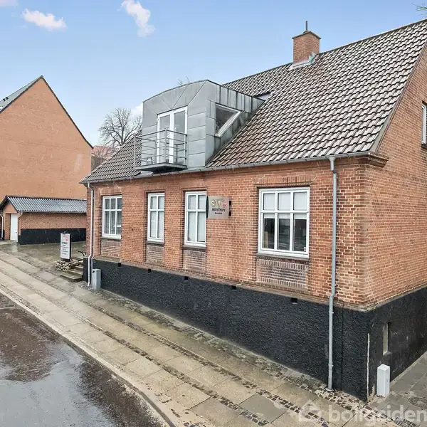 Rødstenshus med skråt tag og tagvinduer ligger på en gade med fortov. Orange bil parkeret ved siden af huset. Huset har flere vinduer og en lille altan.
