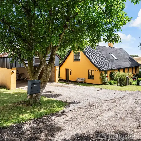 Et gult hus med sort tag står i en frodig have. En grusvej fører op til hovedindgangen omgivet af træer og buske. En garage er placeret ved siden af.