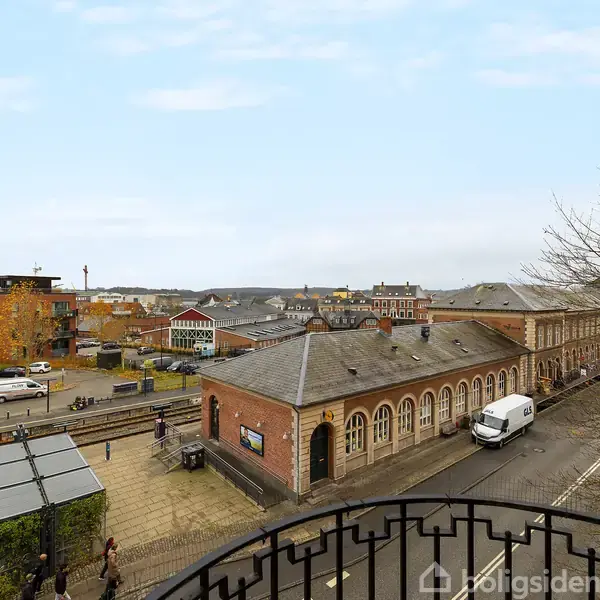 En murstensbygning strækker sig langs en vej, hvor en hvid varevogn er parkeret. I baggrunden ses flere moderne bygninger og jernbanespor under en blå himmel.