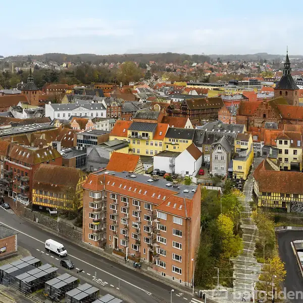 Bygninger med karakteristiske røde tegltage står tæt samlet i en malerisk by, set fra luften. I forgrunden løber en vej med biler, og landskabet strækker sig mod skovklædte bakker i baggrunden.