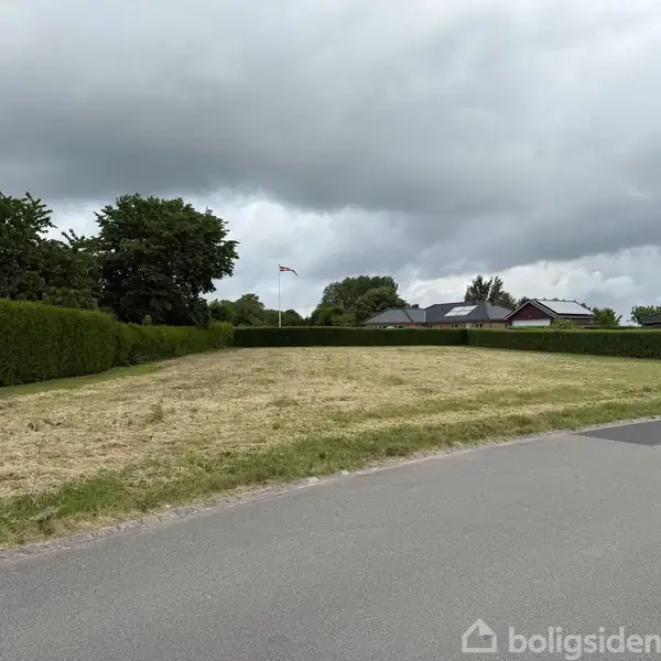 Græsklippen græsplæne omgivet af hække ligger ved en rolig villavej. I baggrunden er der flere huse og en flagstang. Skyet himmel dækker horisonten.