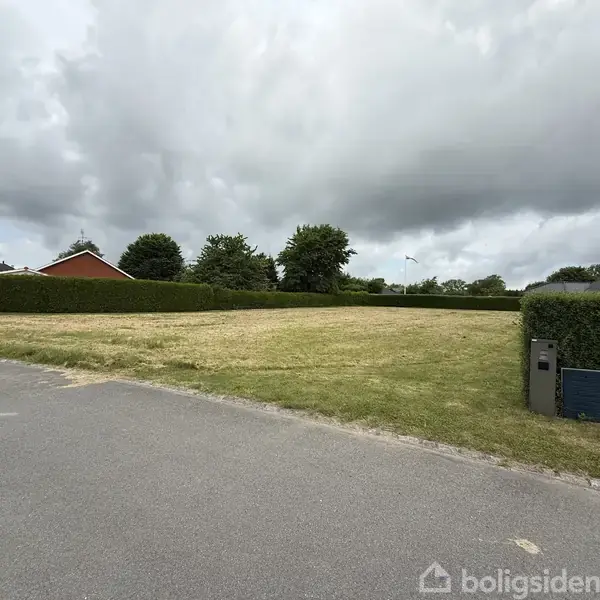 En tom græsplæne strækker sig hen over et parcelhuskvarter omgivet af hække og et par træer i baggrunden under en overskyet himmel.