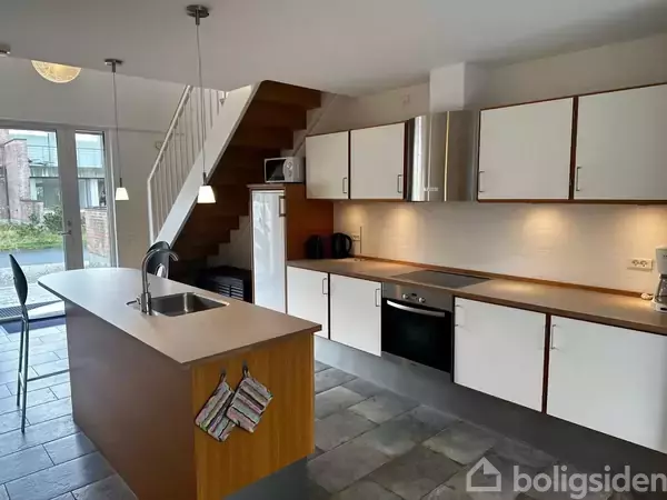 Et moderne køkken med en køkkenø med vask, omgivet af hvide skabe og en ovn. En trappe fører til næste etage, og store vinduer åbner ud til en have.