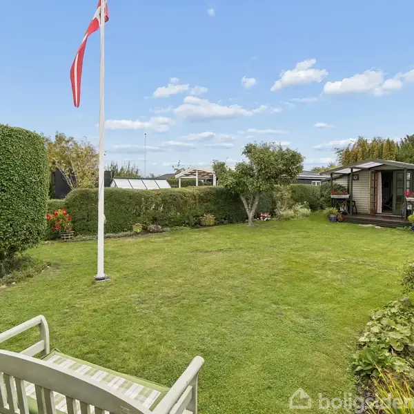 Bænk står på græsplæne foran flagstang med dansk flag, omgivet af trimmede buske og træer. I baggrunden ses en lille bygning. Solrigt vejr og blå himmel med skyer.