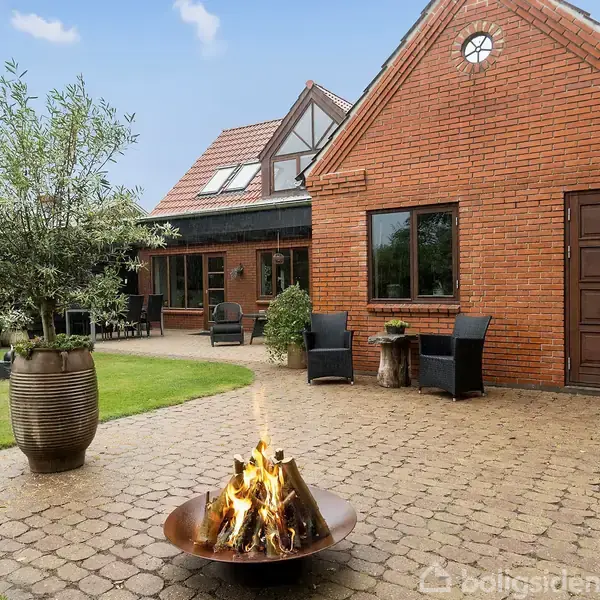Et bål brænder i en ildskål på en flisebelagt terrasse. En rød murstensbygning med brune døre og vinduer omkranser gårdhaven. Plantekrukker og havemøbler pryder terrassen.