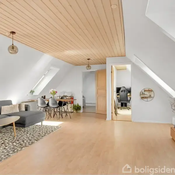 Grå sofa står i stue under skråloft med trægulv og træloft. Fjernsyn på lavt bord, spisebord med stole. Lysindfald fra skråvinduer, indgang til andet rum.